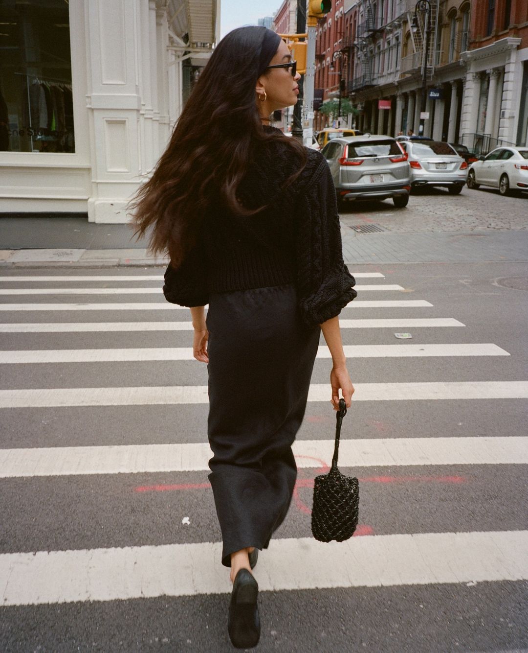 Woman with long dark hair wearing a black knitted cardigan, dark skirt and shoes, walks across a pedestrian crossing in a city.