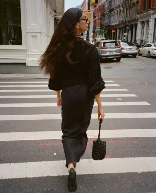Woman with long dark hair wearing a black knitted cardigan, dark skirt and shoes, walks across a pedestrian crossing in a city.