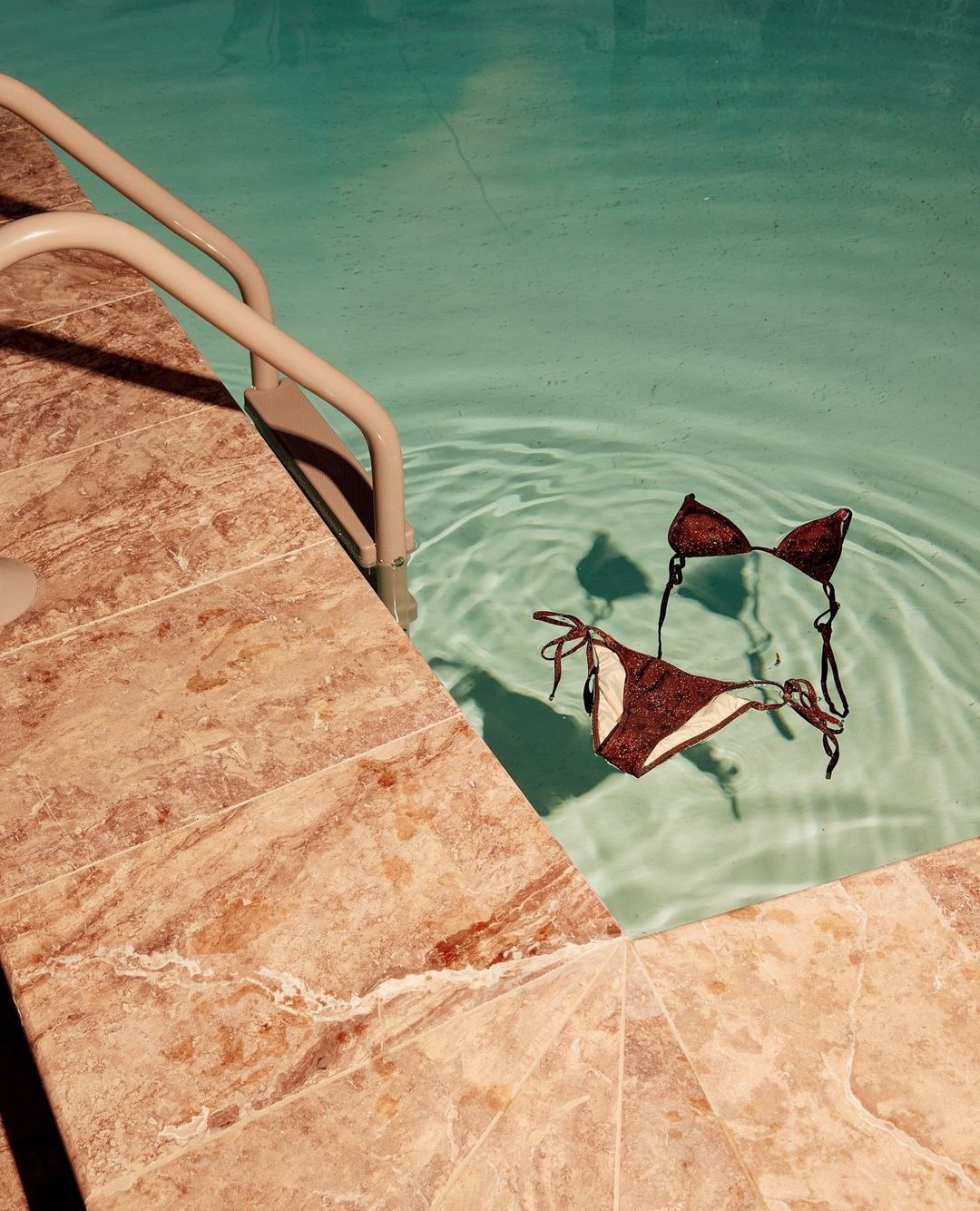 A brown and white bikini floats in a pristine blue swimming pool next to the pool ladder and a tile surround on a sunny day.
