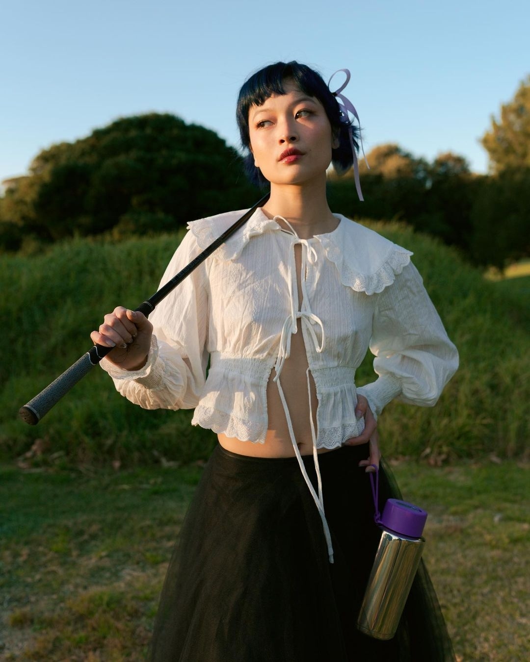 Adult woman in a white top and black skirt poses on a grassy field holding a golf club and a water bottle with trees in the background.