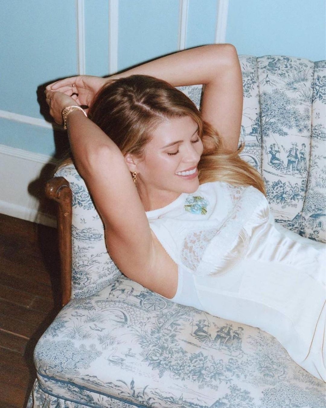 A woman with blond hair is lying on a vintage couch with blue floral patterns, with her hands behind her head, smiling in a casual pose.