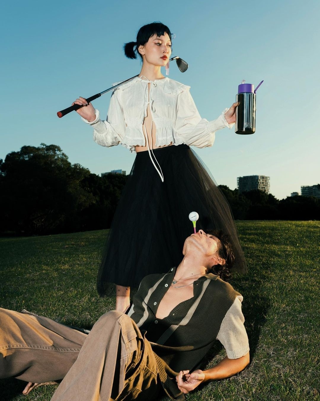 Two people pose outdoors with golf clubs and a water bottle. One person lies on the grass with a golf ball balanced on their face.