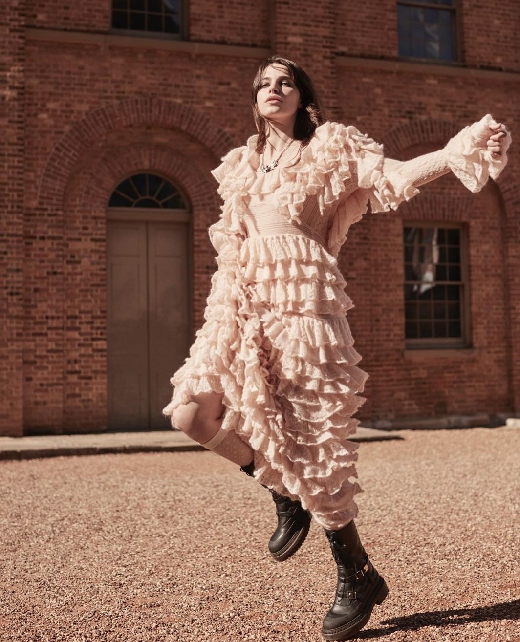 Woman in a ruffled dress and combat boots strikes a pose in an outdoor courtyard with a brick building in the background during daytime.