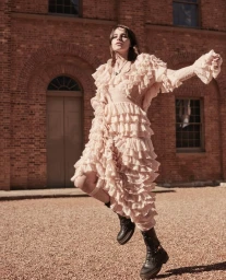 Model in a ruffled beige dress and black boots posing outdoors in front of a red brick building exterior in Sydney.