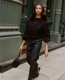 Model in black textured knit sweater and dark skirt walking past green neoclassical columns on a city sidewalk