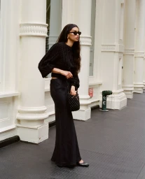 Woman in black sweater and skirt holding a woven handbag while standing outdoors near neoclassical columns