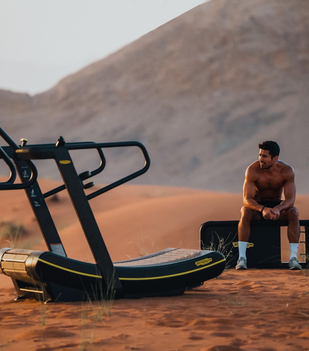Shirtless man rests on an exercise box next to a treadmill in the desert landscape, with sand dunes visible in the background.