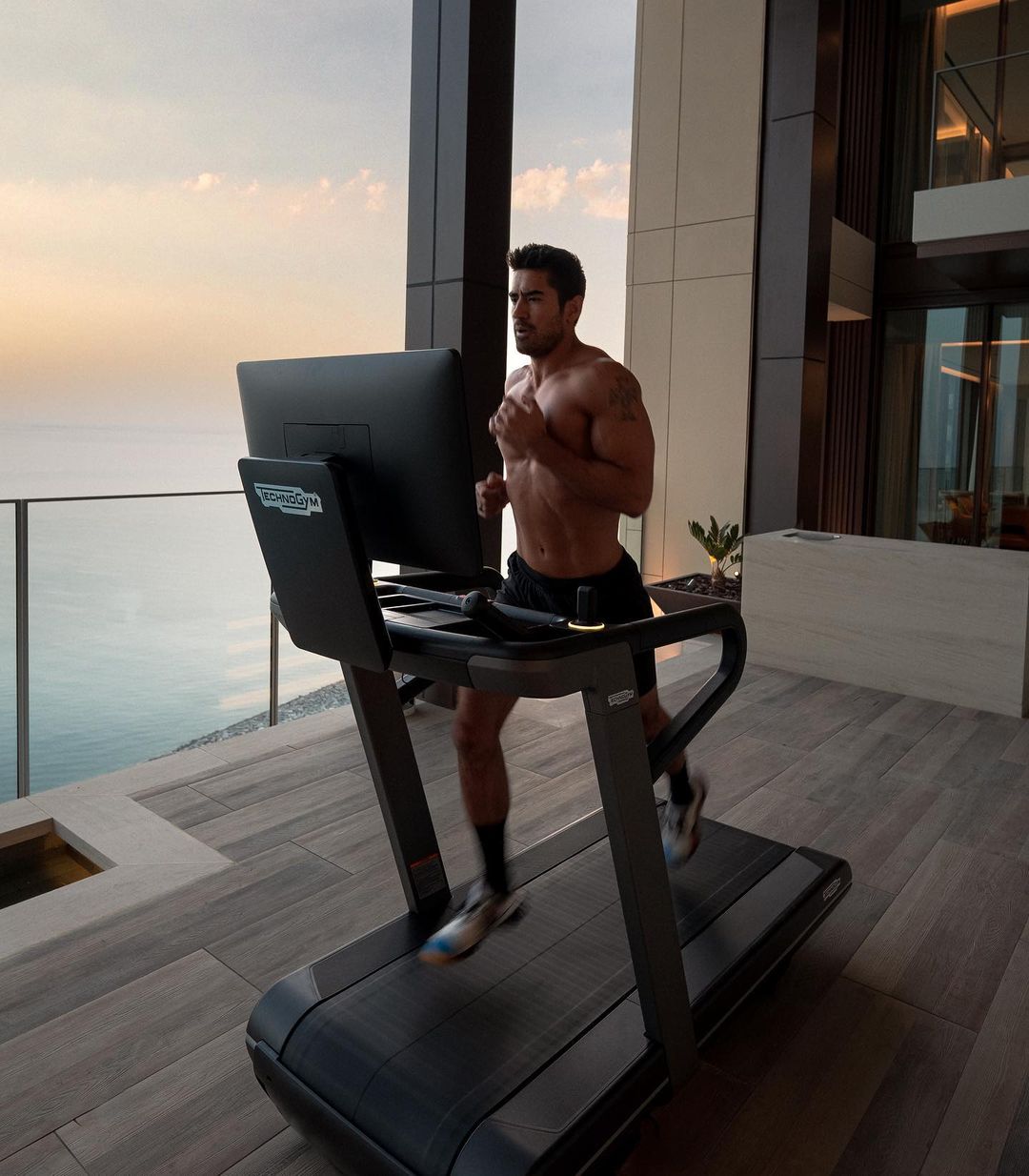 Fit adult male with athletic build runs on a black treadmill inside modern apartment with outdoor ocean view during daytime workout.
