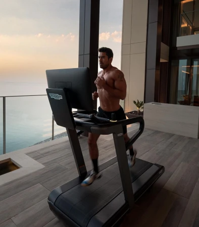 Fit adult male with athletic build runs on a black treadmill inside modern apartment with outdoor ocean view during daytime workout.