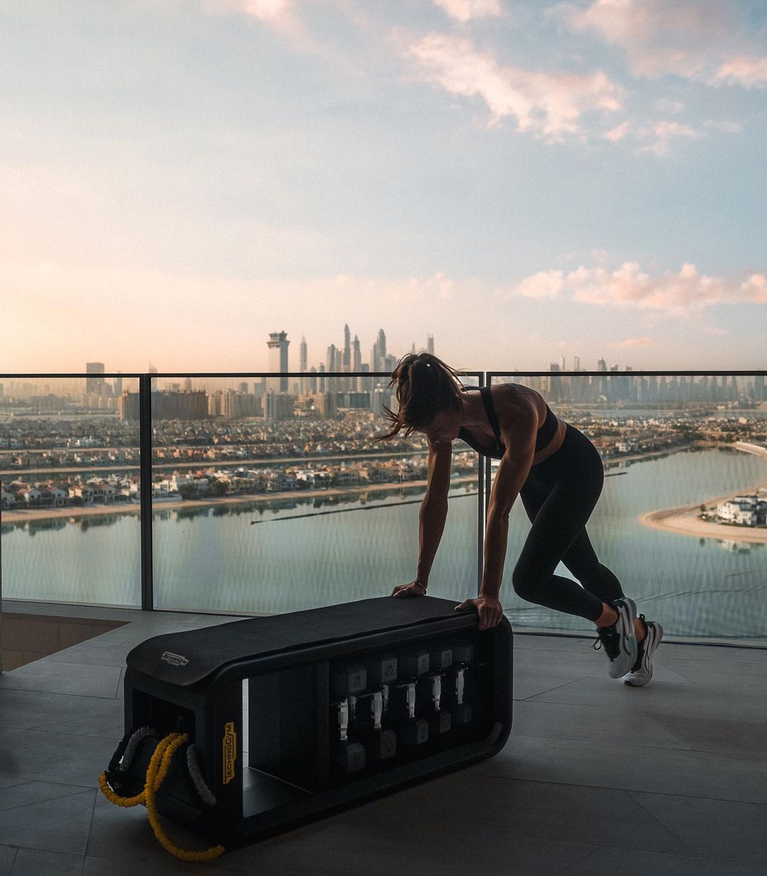 Woman in black athletic wear does step-ups on a workout bench with dumbbells on a rooftop with a city skyline view at sunset.