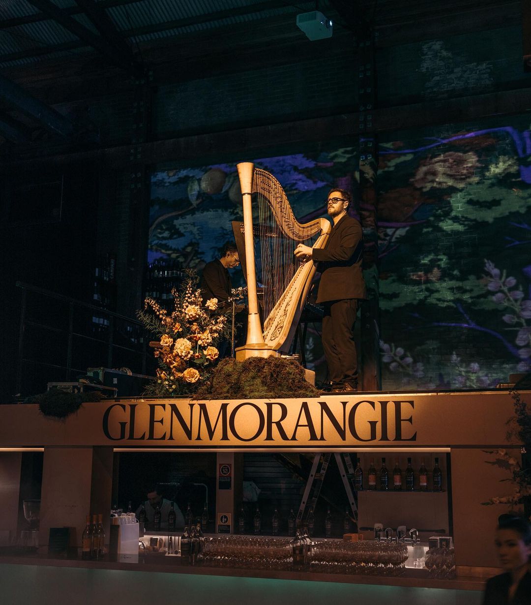 Two musicians perform on an elevated stage with a harp at a Glenmorangie event with floral arrangements and dark, moody lighting.