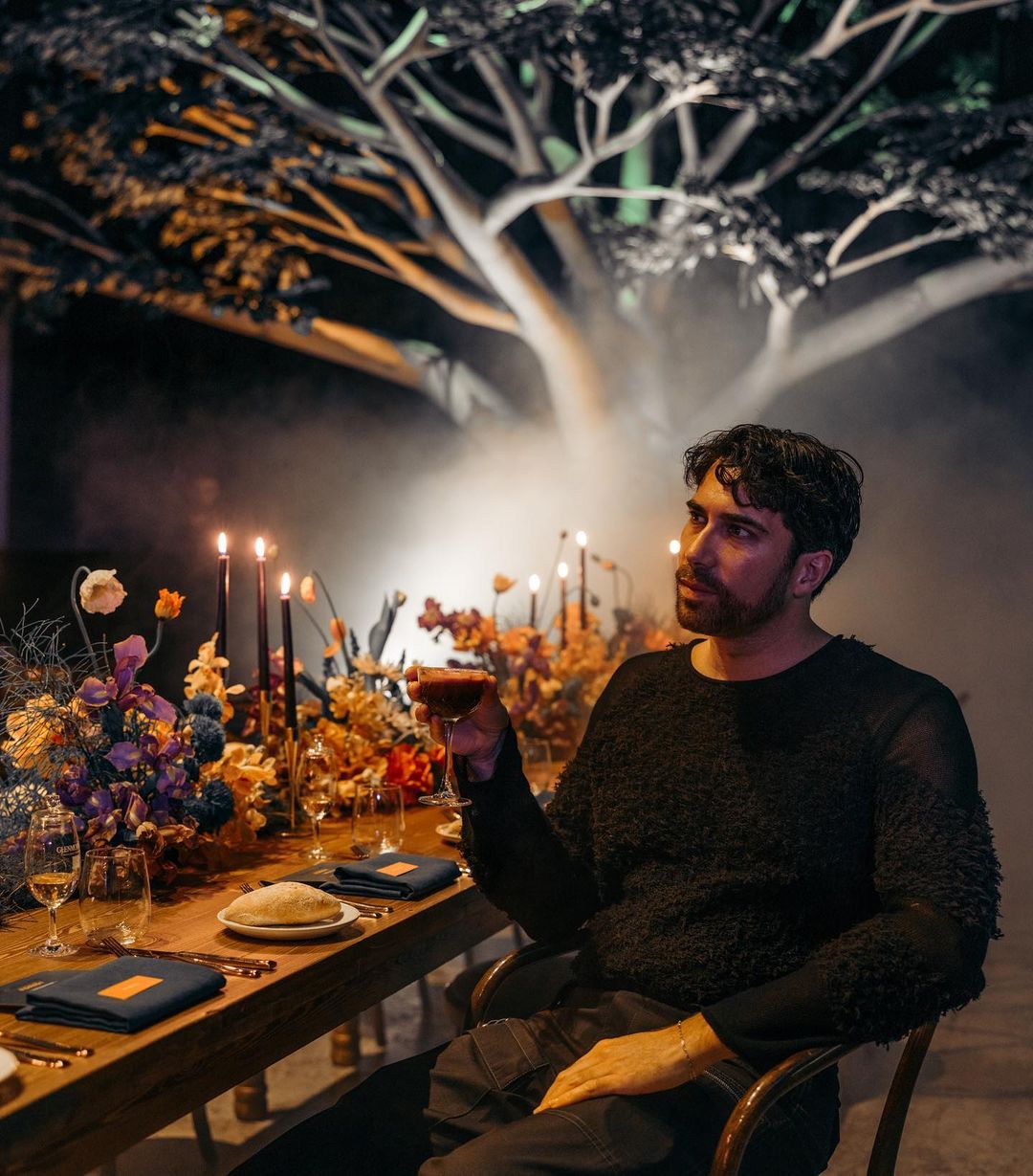 Adult male seated at a table with floral arrangements, candles, glassware, and napkins in an indoor setting with a large tree in the background.