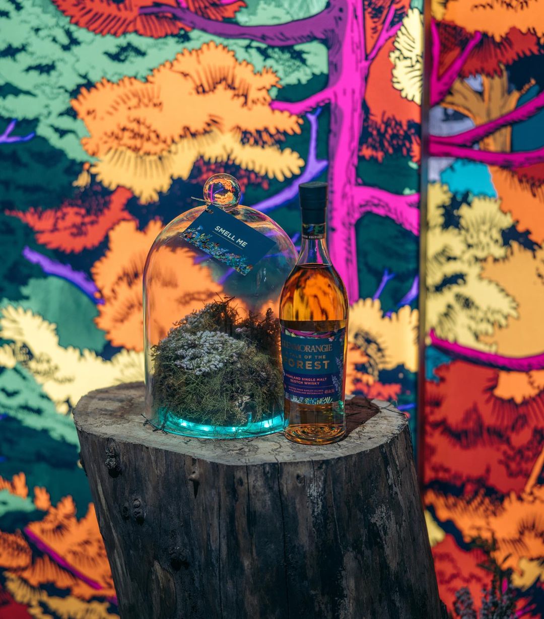 Bottle of single malt scotch whisky beside a dome glass filled with moss on a tree stump with a colorful abstract forest backdrop.