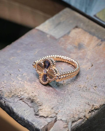 Rose gold ring with four-leaf clover design, featuring a small diamond center, resting on a textured grey surface in a studio setting.