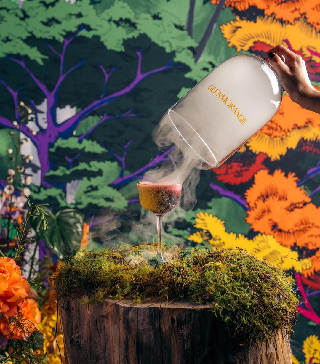 Hand pouring smoky vapor from a Glenmorangie cloche over a cocktail on a mossy tree stump, with a vibrant floral backdrop.