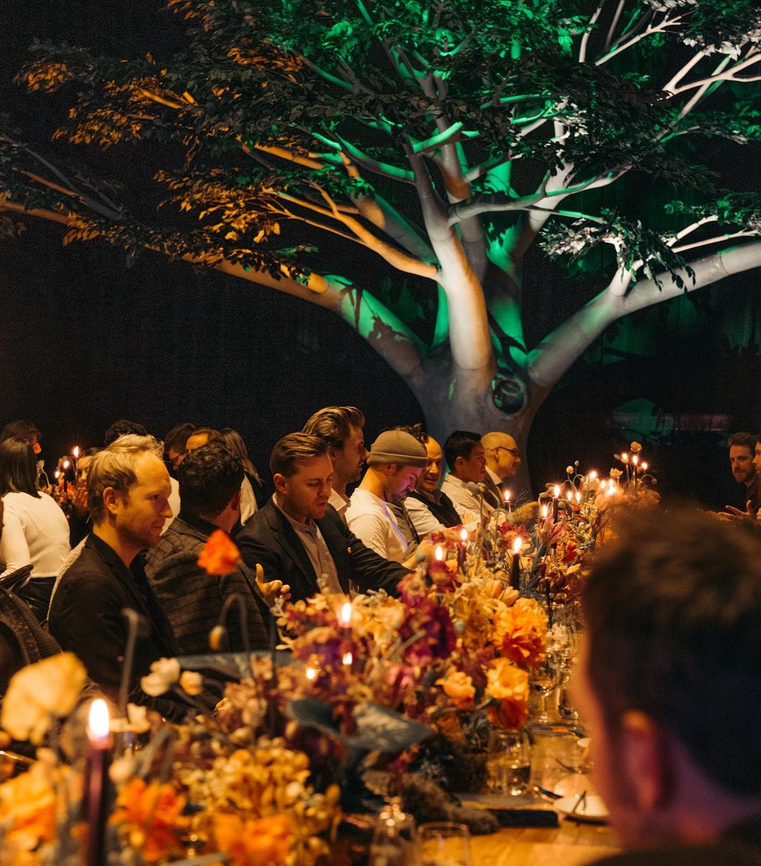 Indoor event with long tables adorned with floral arrangements and lit candles, groups of adult men are seated, and a large tree in background.