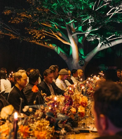 Indoor event with long tables adorned with floral arrangements and lit candles, groups of adult men are seated, and a large tree in background.