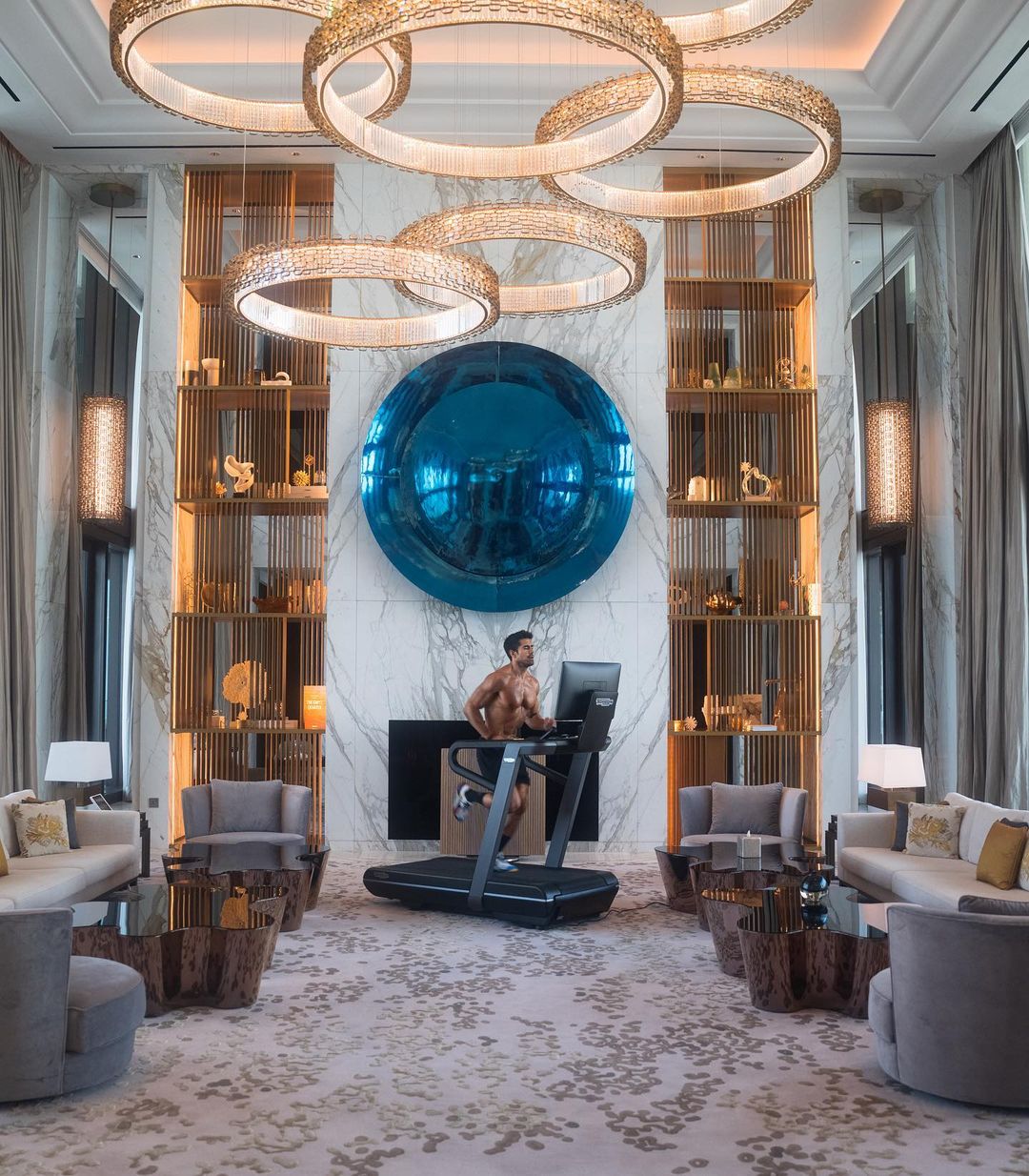 Bare-chested man uses a treadmill indoors; luxury lounge setting with multiple sofas, round chandeliers, and a large blue spherical wall decor.