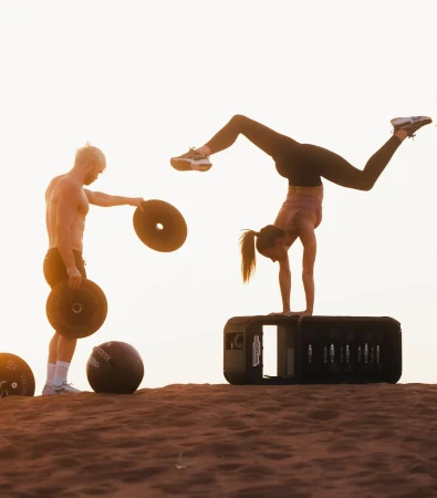 Female athlete doing a handstand on a box as a man spotter holds weight plates, outdoors at sunset, athletic gear and sportswear visible.