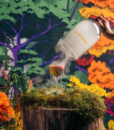 A hand pours smoke from a glass cloche over a cocktail resting on a moss-covered tree stump against a colorful graphic wallpaper backdrop