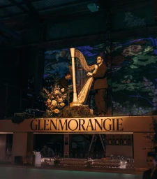 Musician playing a harp on an elevated stage featuring large illuminated Glenmorangie branding and floral arrangements.