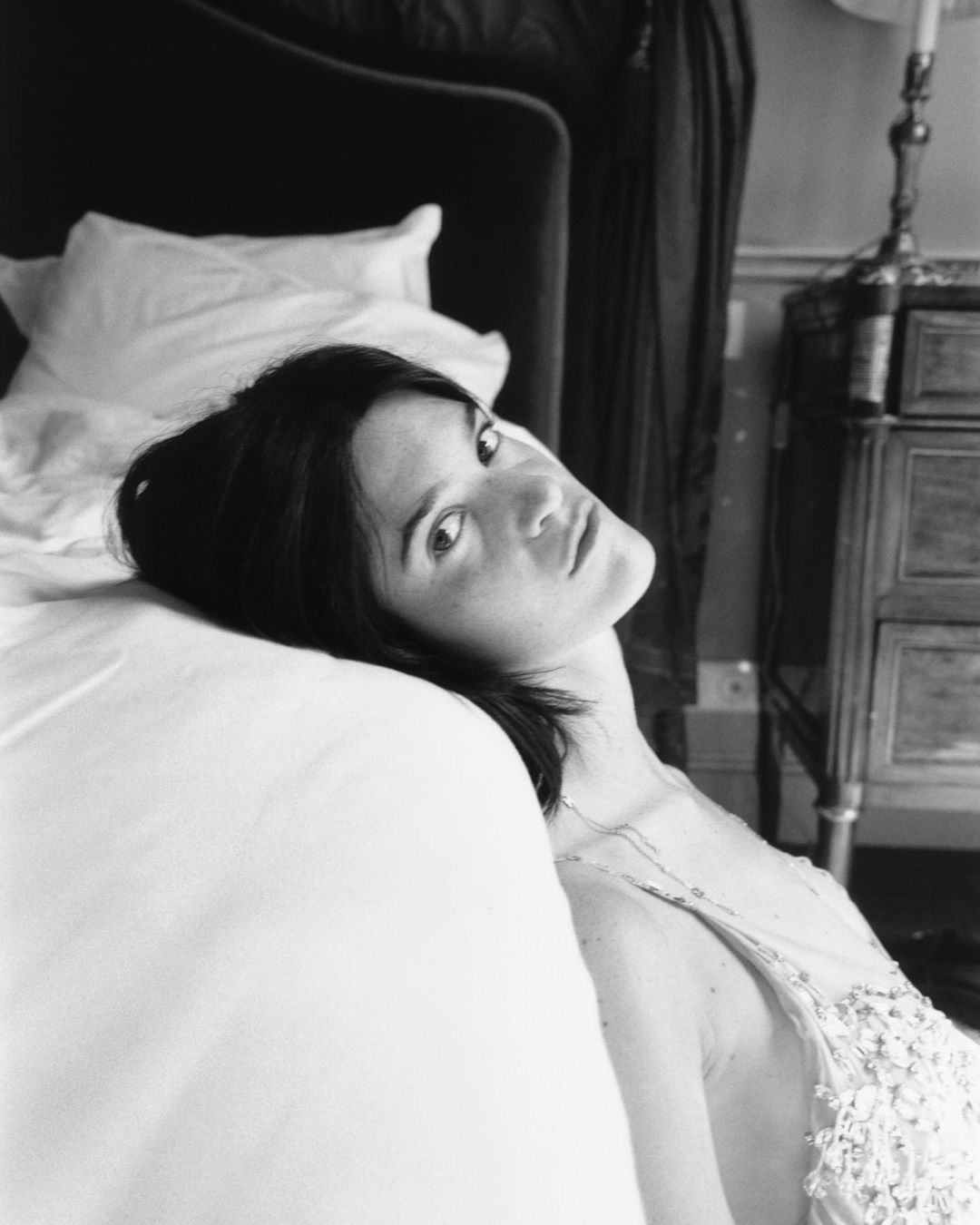 Black and white portrait of a woman lying on a white bed with a vintage bedside table and lamp, shot with natural lighting.