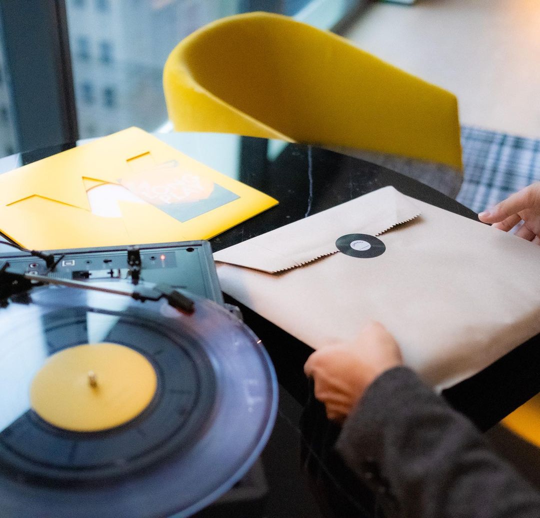 Close-up of a person placing a record album in a brown paper sleeve, set on a black marble table with a turntable and yellow chair.
