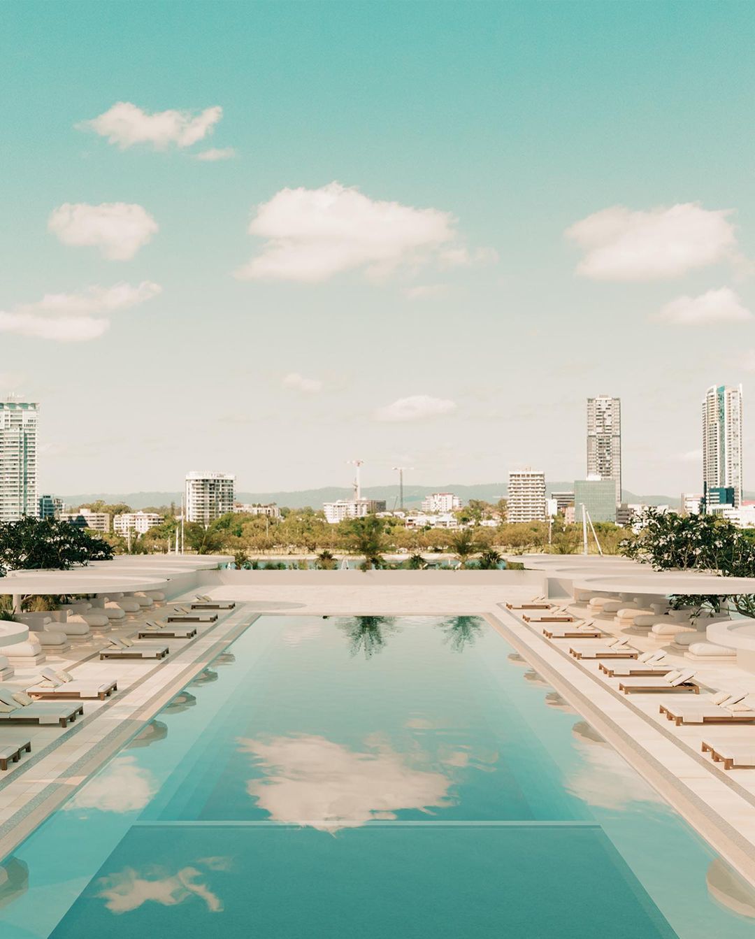 Rooftop pool reflects sky and clouds with a city skyline in the background, lounge chairs on the pool deck, creating a serene urban oasis.
