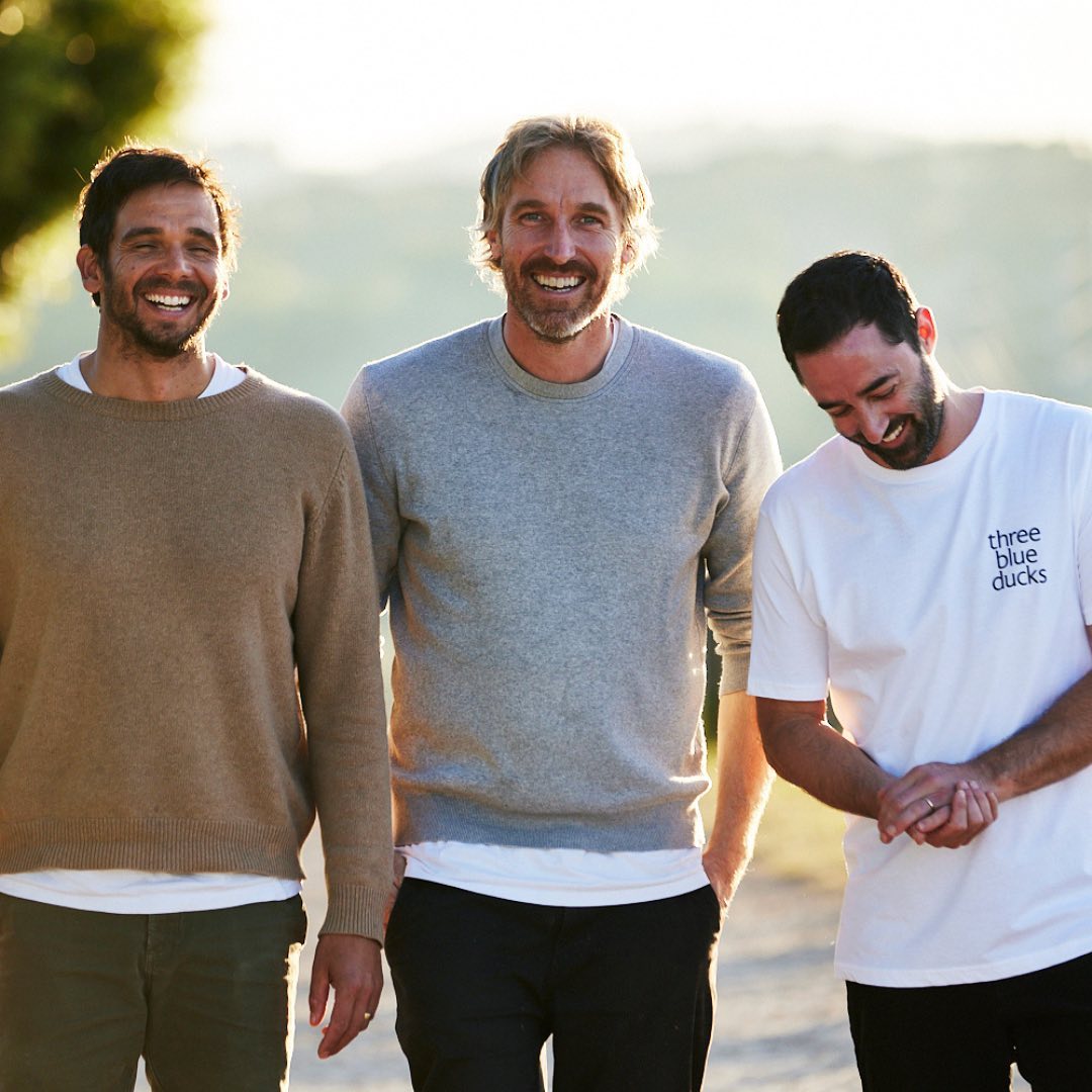 Three adult men stand together outdoors in casual clothing, smiling in a medium shot with a blurred background, enjoying natural light.