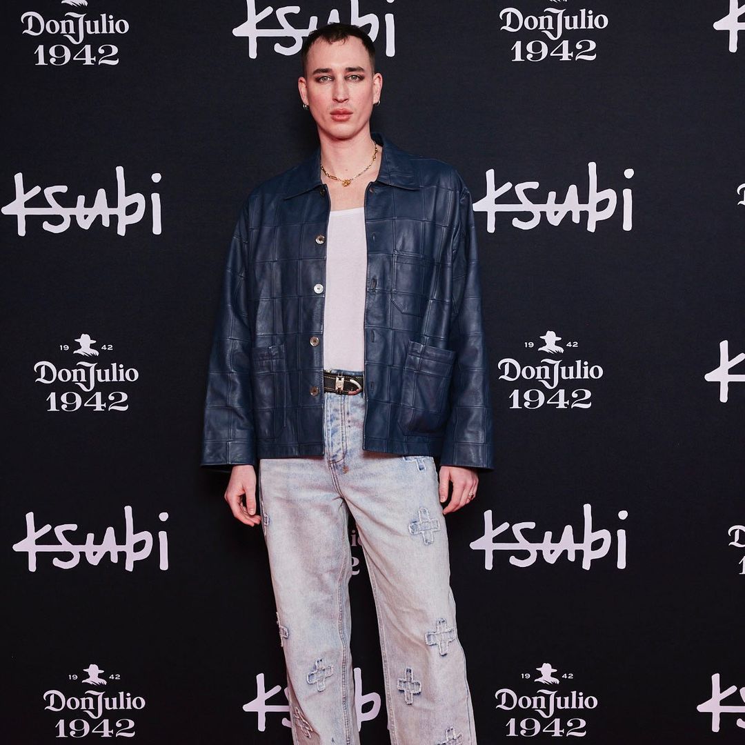 Adult in a blue leather jacket and light-wash jeans with cross details, poses in front of a branded backdrop, captured in a medium shot.