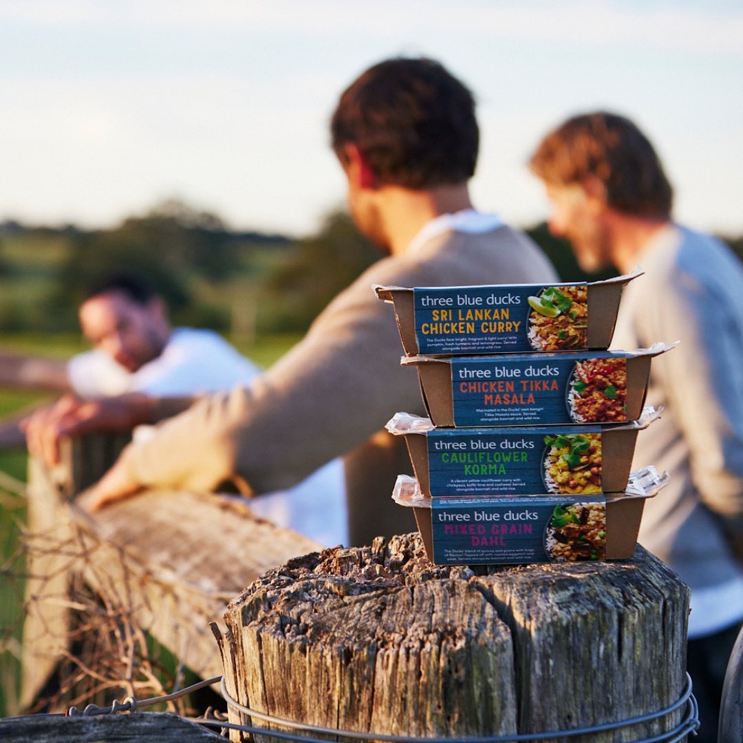 Stack of Three Blue Ducks meal containers on a wooden post outdoors, with three men visible in the background in a rural setting.