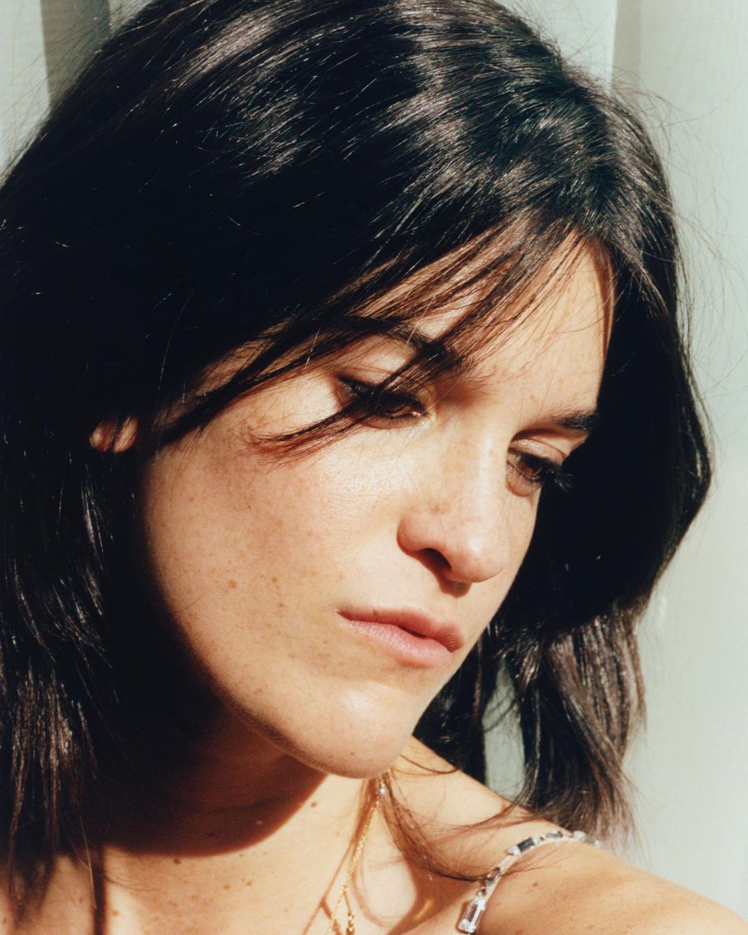Close-up portrait of a young adult woman with dark hair and fair skin, gazing downward in soft lighting, shot with shallow depth of field.