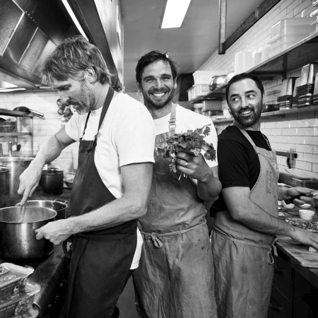 Three chefs work in a commercial kitchen, preparing food at stainless steel counters, with one holding a bunch of cilantro.