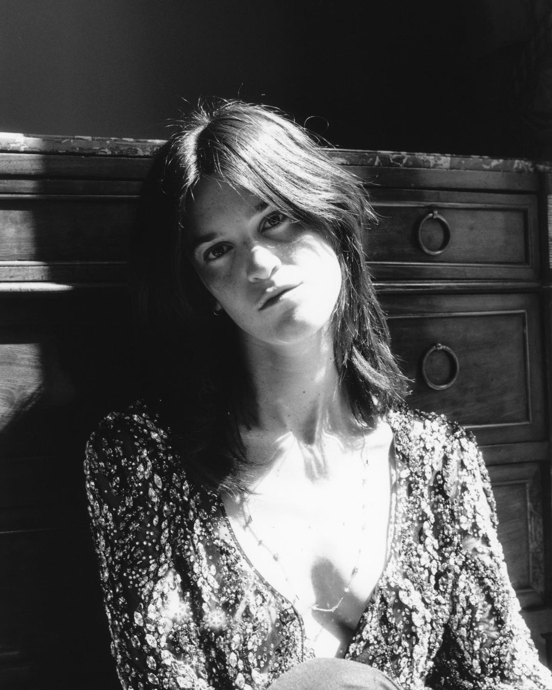 Monochrome portrait of a young adult woman with shoulder-length hair wearing a patterned top, seated in front of a wooden cabinet.