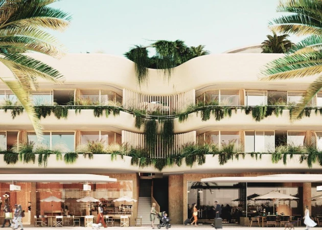 Exterior of a modern multi-story building with balconies and abundant greenery, people strolling in front, palms, and clear sky.