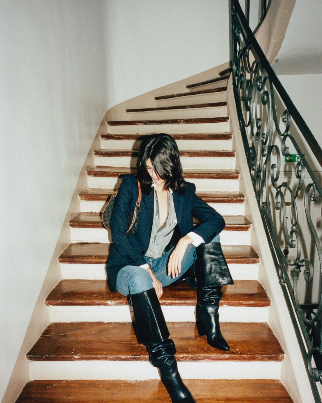 Brunette woman wearing a navy blazer, jeans, and tall black boots sitting on wooden steps indoors near a black spiral staircase.