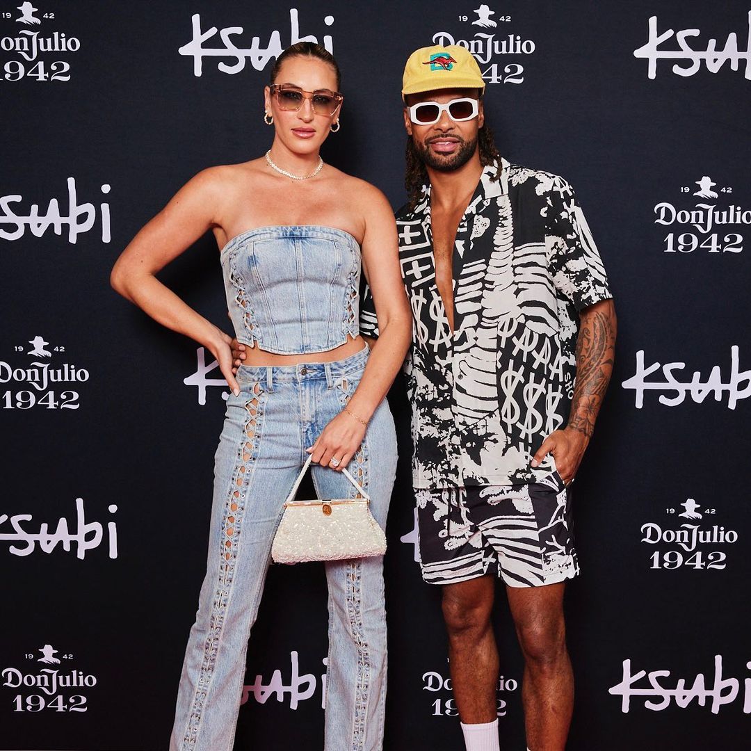 Two fashionably dressed people, a woman in denim and a man in a patterned shirt and shorts, pose in front of a branded backdrop.