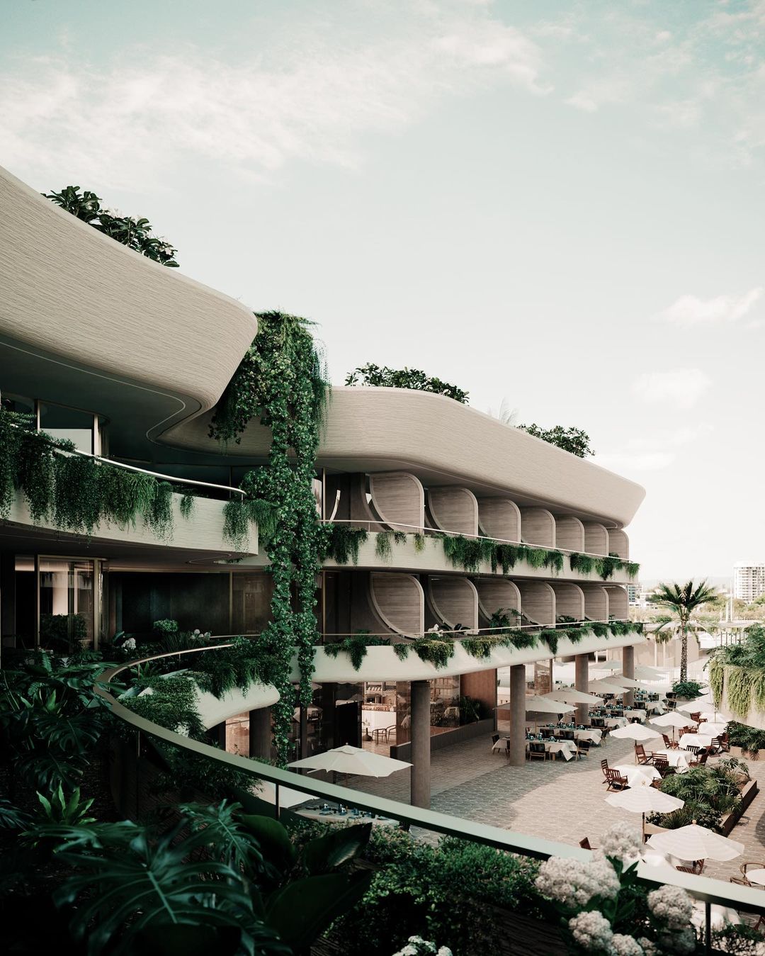 Exterior view of a multi-story hotel with curved balconies, lush greenery, outdoor dining area, and umbrellas on a sunny day.