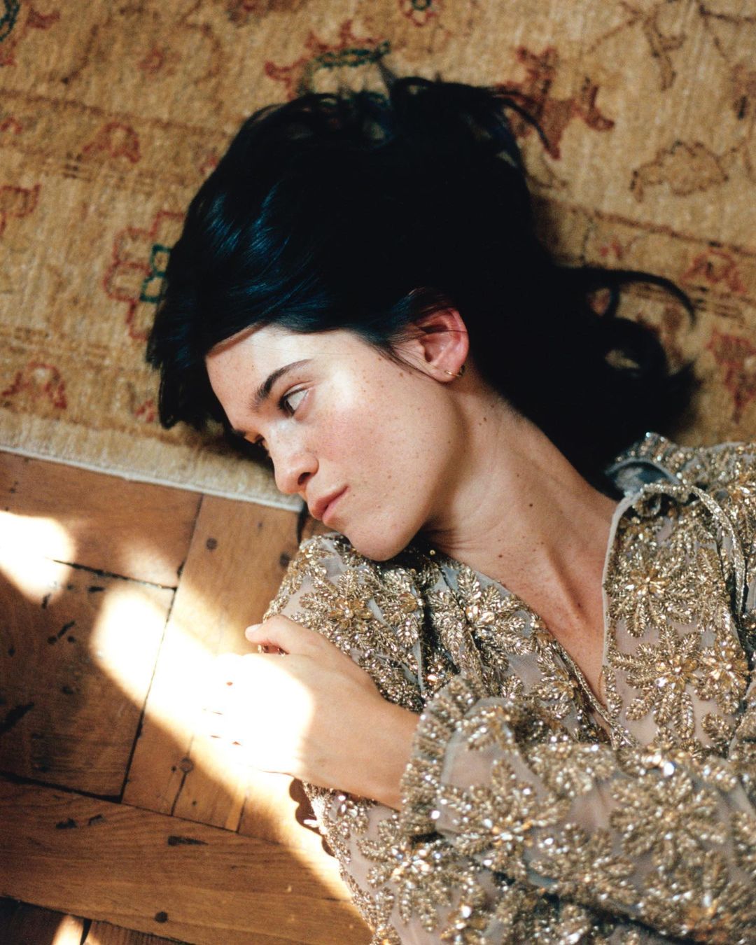 Brunette woman with fair skin lying on a carpeted floor with light shining on wooden floorboards and a patterned, beaded shirt.