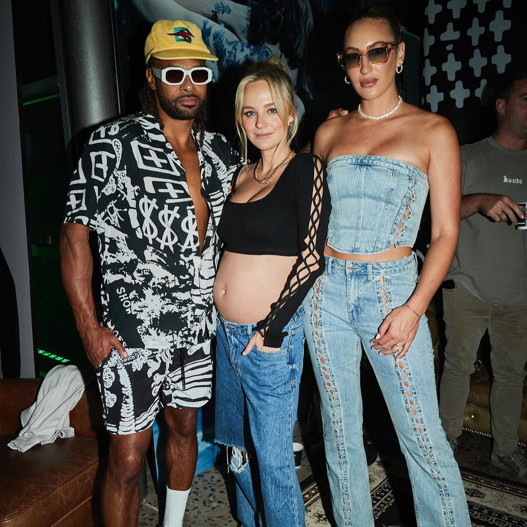 Three people pose indoors at an event, featuring two women in denim outfits and a man in a patterned shirt and shorts.