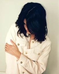 Woman with dark hair wearing a cream jacket with gold buttons against a neutral wall