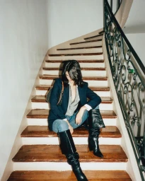 Woman in dark blazer and black boots seated on a wooden staircase with ornate iron railing