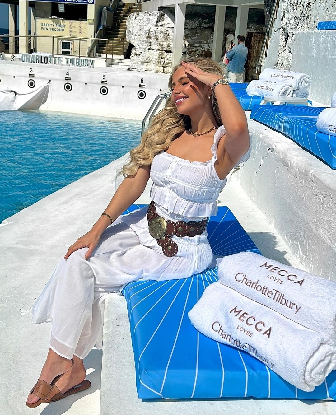 A blonde woman in a white dress smiles while sitting on a blue lounger next to a pool with towels, capturing a serene moment.