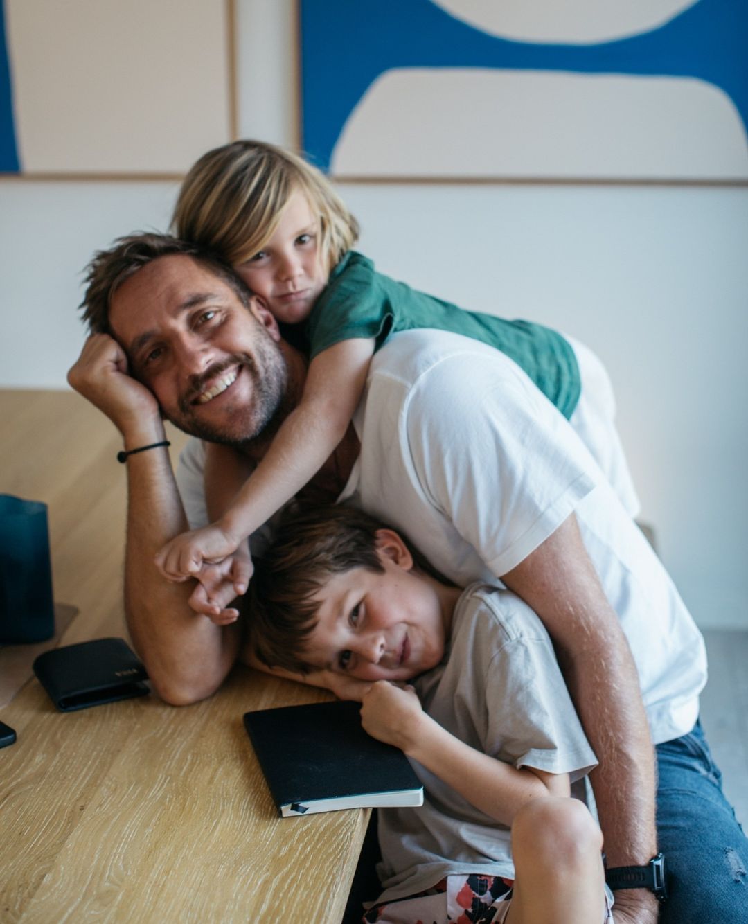 Father and two children pose at a wooden table indoors in a medium shot. The father smiles, and the kids are on top of him.
