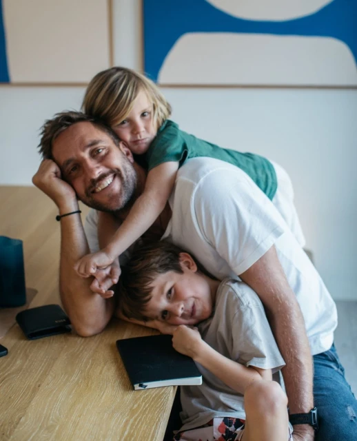 Father and two children pose at a wooden table indoors in a medium shot. The father smiles, and the kids are on top of him.