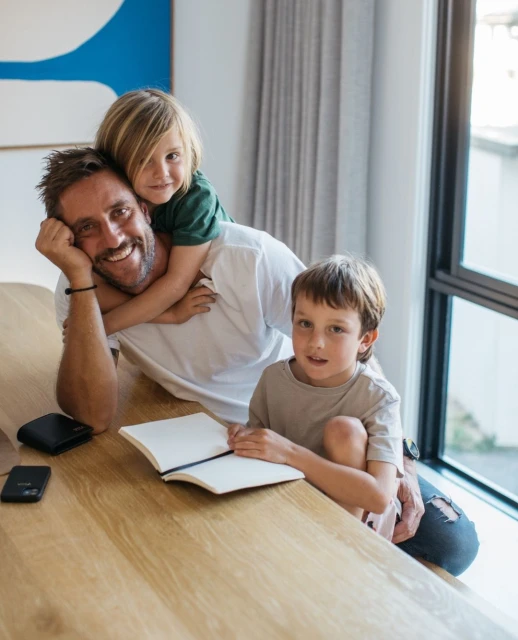 A smiling dad sits at a wooden table with two children, one on his back and one sitting beside him, in a bright, modern home setting.
