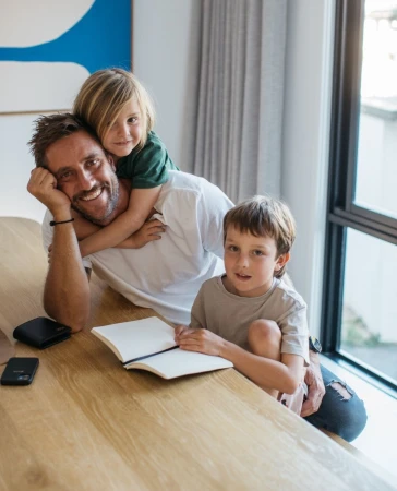 A smiling dad sits at a wooden table with two children, one on his back and one sitting beside him, in a bright, modern home setting.