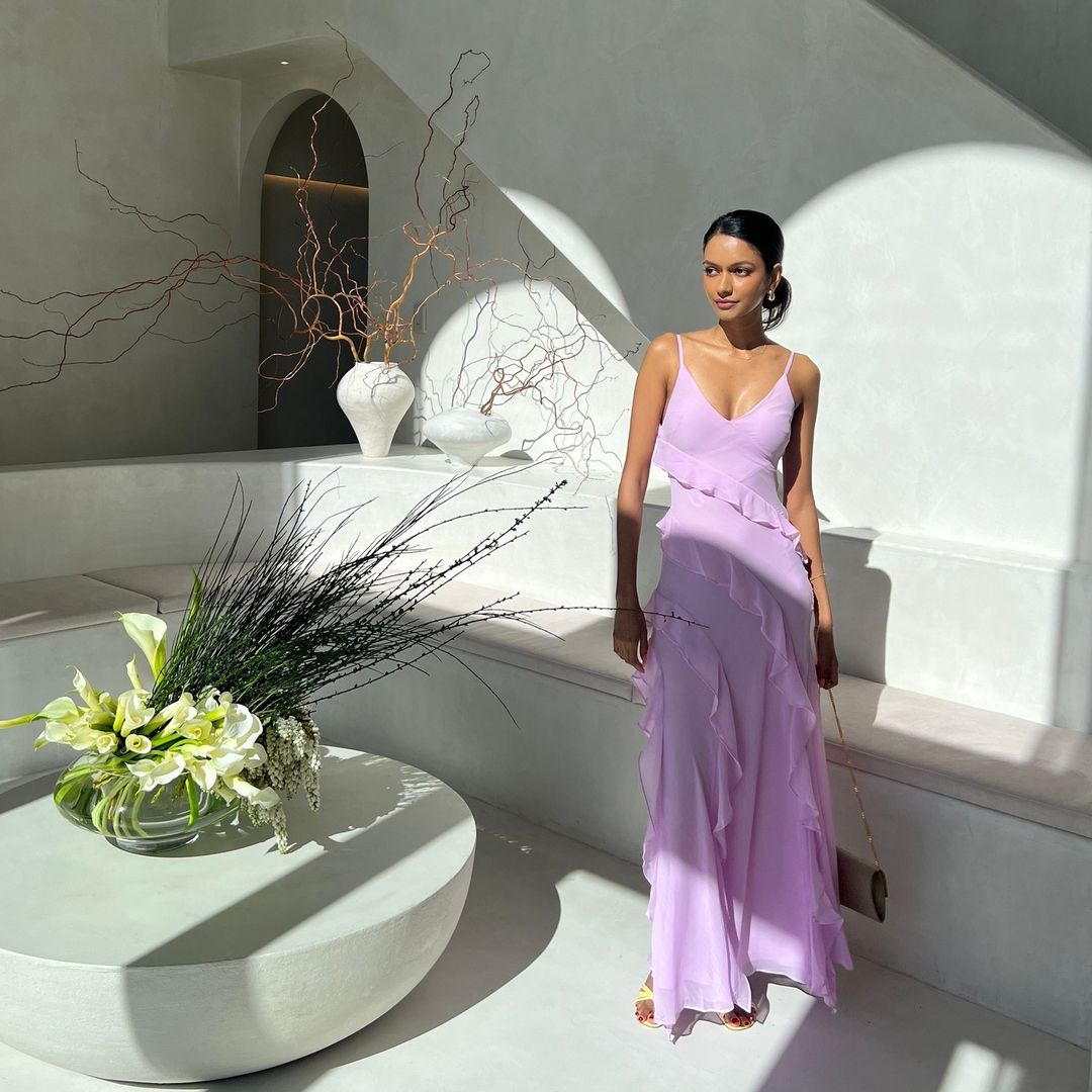 Woman in a lavender gown stands in a brightly lit interior with modern architectural details and a white flower arrangement.