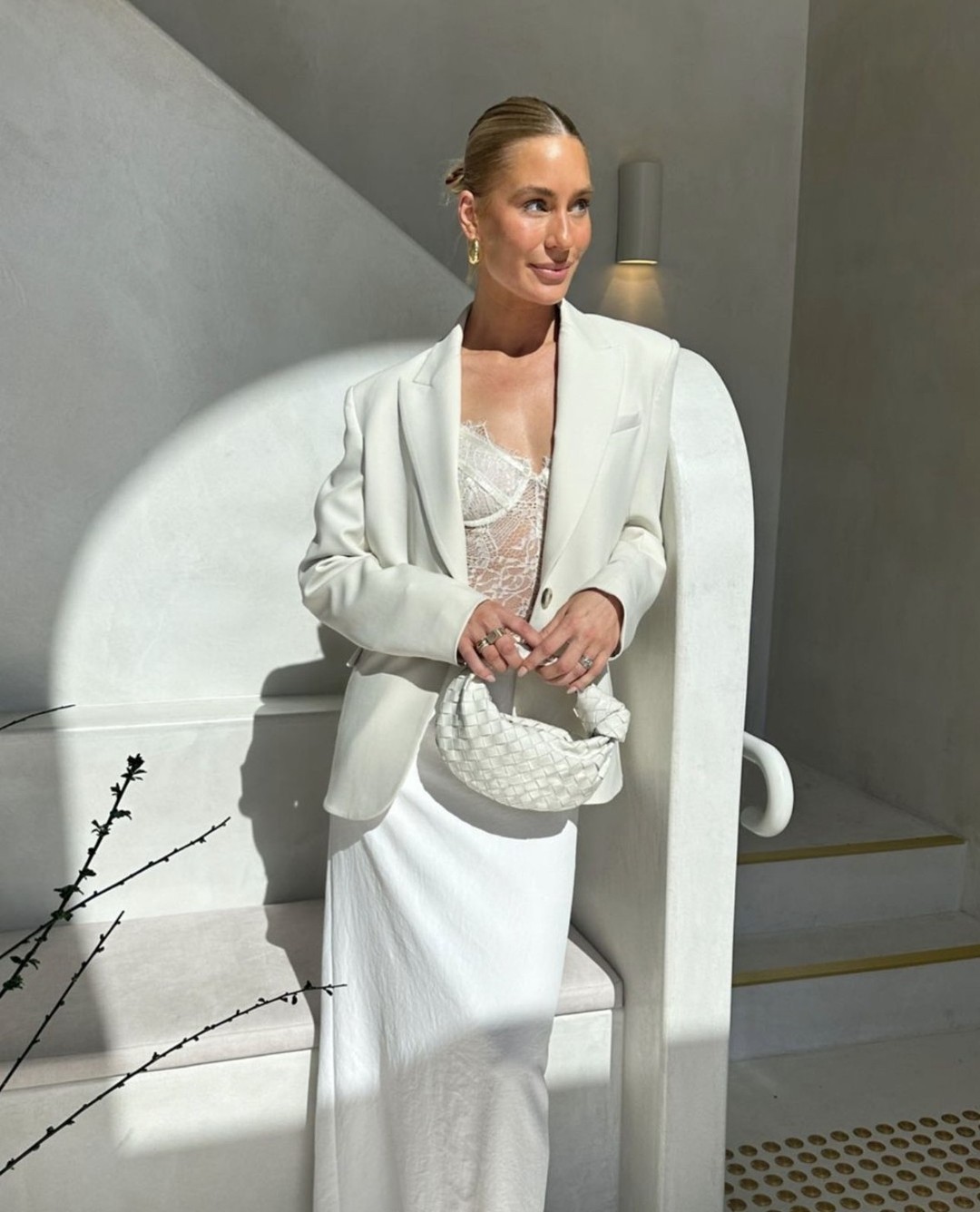 A woman with blonde hair, wearing a white blazer, a lacy top, and a white skirt, poses with a woven bag indoors by white architectural elements.