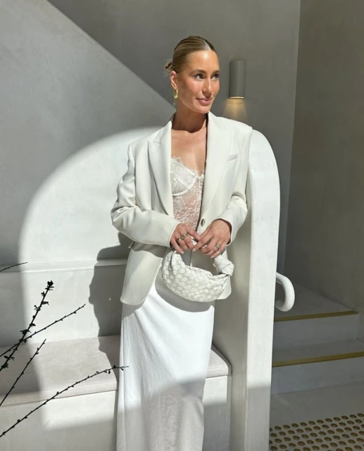 A woman with blonde hair, wearing a white blazer, a lacy top, and a white skirt, poses with a woven bag indoors by white architectural elements.